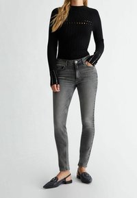 Top largo de manga larga negro con detalles cortados, combinado con jeans ajustados gris claro y zapatos planos negros con embellishments.