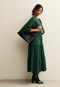 Grünes Kleid mit Stufen und strukturierter Stoffqualität, kombiniert mit einer schwarzen Schultertasche. Modell trägt schwarze Stiefel mit Absatz. Schlichter Hintergrund.