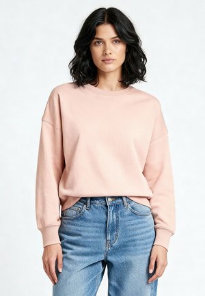 Sweat-shirt rose clair avec un col rond et des épaules tombantes, fabriqué en tissu doux. Associé à un jean bleu taille haute.