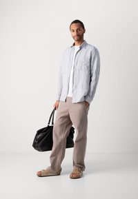 Light blue striped button-up shirt over a white t-shirt, beige pants, and light sandals; holding a black duffle bag.