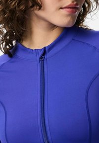 Ljubičasti sportski top s visokim ovratnikom i kratkim zatvaračem. Izrađen od glatkog materijala, s kontrastnim šavovima i prilagođenim krojem.