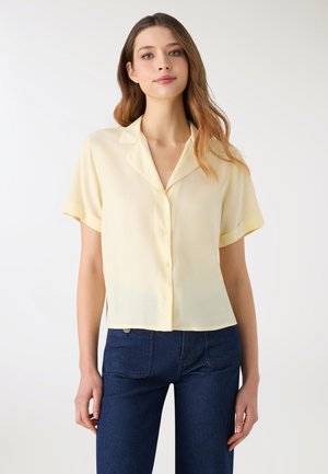 Krótka żółta bluzka z kołnierzykiem, zapięciem na guziki z przodu oraz lekką, przezroczystą fakturą. W połączeniu z granatowymi spodniami dżinsowymi.
