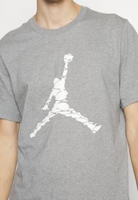Gros plan sur une personne portant une chemise grise avec une silhouette graphique blanche d'une figure sautant, composée de formes irrégulières, sur la poitrine.