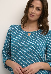 Bluză teal cu un model ondulat alb, având un decolteu rotund și o mică deschidere în față. Mâneci ¾ cu o croială lejeră. Textură a materialului moale.