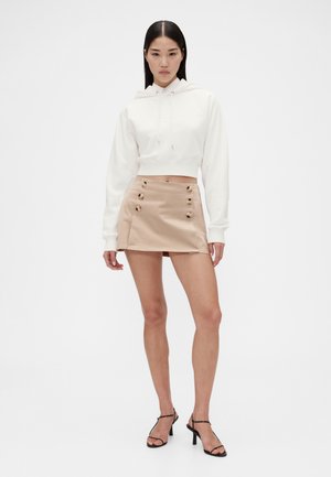 Witte cropped hoodie met borduursel aan de voorkant, gecombineerd met een beige minirok met zes knoopdetails en een gladde textuur. Zwarte sandalen met bandjes.