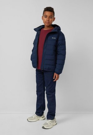 Marineblauer gepolsterter Mantel mit Kapuze und Reißverschluss, mit einem Logo auf der Brust. Kombiniert mit einem burgunderfarbenen Hemd und dunkelblauen Jeans, vervollständigt mit Sneakers.