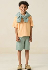 T-shirt à manches courtes orange clair, shorts vert menthe avec poches latérales, pull vert menthe noué au cou, chaussures sans lacets beiges.