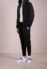 Felpa nera con zip e cappuccio grande, abbinata a pantaloni jogger neri e sneakers bianche con lacci neri. Tessuto morbido, vestibilità rilassata.
