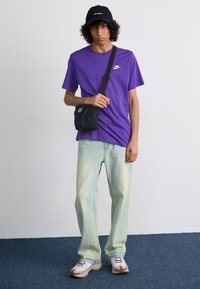 Lilla kortærmet skjorte med Nike-logo, lyseblå loose-fit jeans, sort kasket og en lille sort crossbody taske. Hvide sko med brune detaljer.