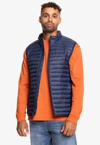 Chaleco acolchado azul marino con cuello alto, cierre de cremallera y sin mangas, superpuesto sobre una camiseta de manga larga naranja, combinado con jeans azules.