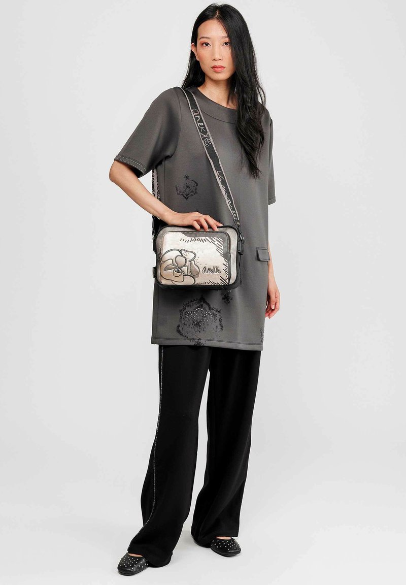 Túnica gris de gran tamaño con acentos texturizados, combinada con pantalones anchos negros. Presenta un bolso cruzado plateado con un diseño floral y strap negro.