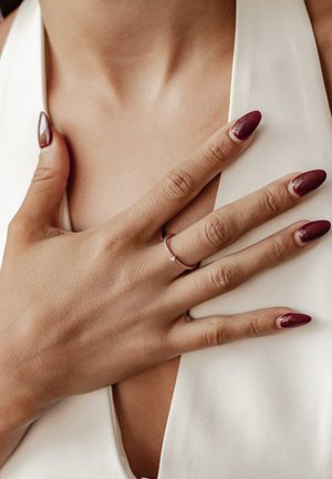 Rosa Goldring mit kleinen Edelsteinen, getragen am Ringfinger. Die Hand hat manikürte Nägel in einem tiefen Burgunderrot und ist vor einem weißen Oberteil platziert.