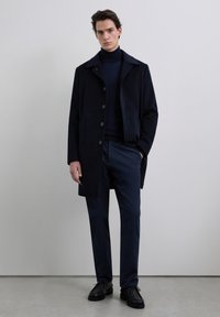 Abrigo de mezcla de lana azul marino con cuello de solapa y seis botones, combinado con un cuello alto a juego y pantalones de corte recto, complementado con zapatos negros.