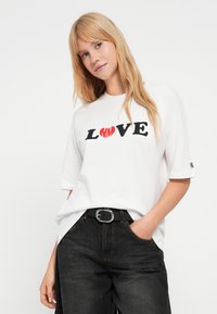 Λευκό υπερμεγέθες t-shirt με μαύρο και κόκκινο γράφημα "LOVE". Το μοντέλο φοράει μαύρο τζιν με ασημένια αγκράφα ζώνης, τα χέρια στις τσέπες.