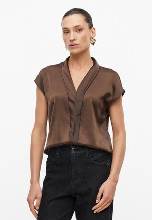 Vrouw draagt een bruine satijnen korte-mouwen blouse ingestopt in een hooggetailleerde zwarte jeans, geaccentueerd met grote gouden oorbellen.