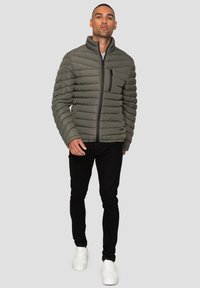 Gepolsterte olivgrüne Zip-Jacke mit Stehkragen, horizontalem Steppdesign und einer schwarzen Reißverschlusstasche auf der Brust. Getragen mit schwarzen Hosen und weißen Sneakers.