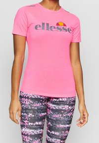 Camiseta deportiva rosa de manga corta con el logo gris de "ellesse", detalles en los hombros y un diseño ajustado. Se lleva con leggings estampados.