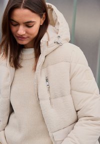 Doudoune beige avec une texture en polaire, poche zippée et capuche réglable. Portée par-dessus un pull en tricot crème clair.