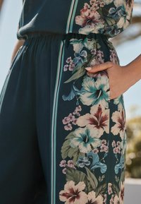 Bloemen jumpsuit in donkergroene stof met grote hibiscussen en kleinere bloemenpatronen, met strepen aan de zijkant en een gegatherde taille.