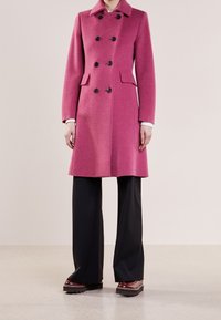 Roze dubbelzijdige jas met een kraag, zwarte knopen, zijzakken en lange mouwen, gecombineerd met zwarte wijde broekspijpen en bruine schoenen.