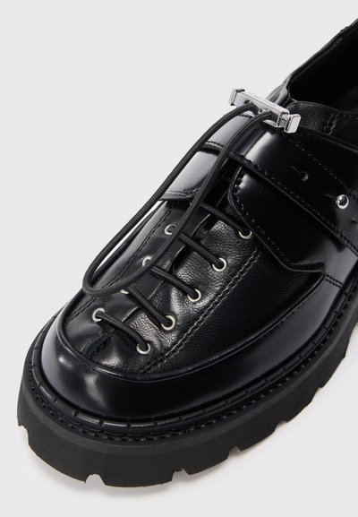 Dettaglio ravvicinato di una scarpa in pelle nera con suola in gomma spessa, lacci elastici senza nodo fissati da una fibbia metallica e dettagli cuciti.