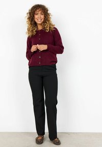 Donna con capelli ricci che indossa un cardigan buttonato bordeaux, pantaloni neri e ballerine marroni con borchie, in piedi davanti a uno sfondo bianco.
