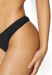 Schwarze Bikinihose mit glattem Stofffinish, niedrig geschnittenem Design und minimaler Abdeckung, ergänzt durch subtile Nähdetails.