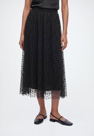 Femme portant une jupe midi en tulle noire à pois et des chaussures plates Mary Jane noires, debout devant un fond uni.