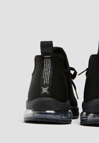 Černé síťované tenisky mají texturovaný svršek, polstrovaný límec a průhlednou vzduchovou odpruženou podrážku. Na patě je značení a QR kód.