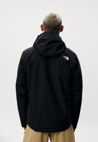 Veste imperméable noire avec capuche, arborant un logo sur l'épaule gauche et une coupe droite. Texture lisse et style minimaliste.