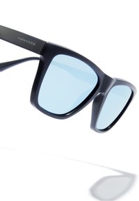 Fekete napszemüveg kék lencsékkel, merész, négyzet alakú kerettel. A szárak vékonyak, matt felületűek, és az egyik oldalon "HAWKERS" márkajelzés található.