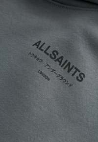 Tessuto grigio con il nome del marchio "ALLSAINTS" stampato in nero, con ulteriore testo in giapponese e "LONDON" sottostante. Design casual.
