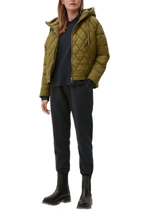 Veste matelassée vert olive avec capuche, associée à un sweat-shirt sombre et un pantalon de jogging noir. Bottines noires à semelles épaisses.