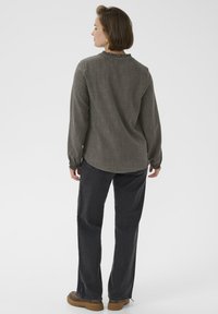 Chemise à manches longues vert olive avec un col volanté, associée à un jean en denim foncé et des chaussures à plateforme brunes, vue de dos.