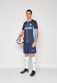 Camisola de futebol azul marinho e turquesa com logotipo, acompanhada de calções azul marinho, meias brancas e chuteiras pretas, segurando uma bola de futebol.
