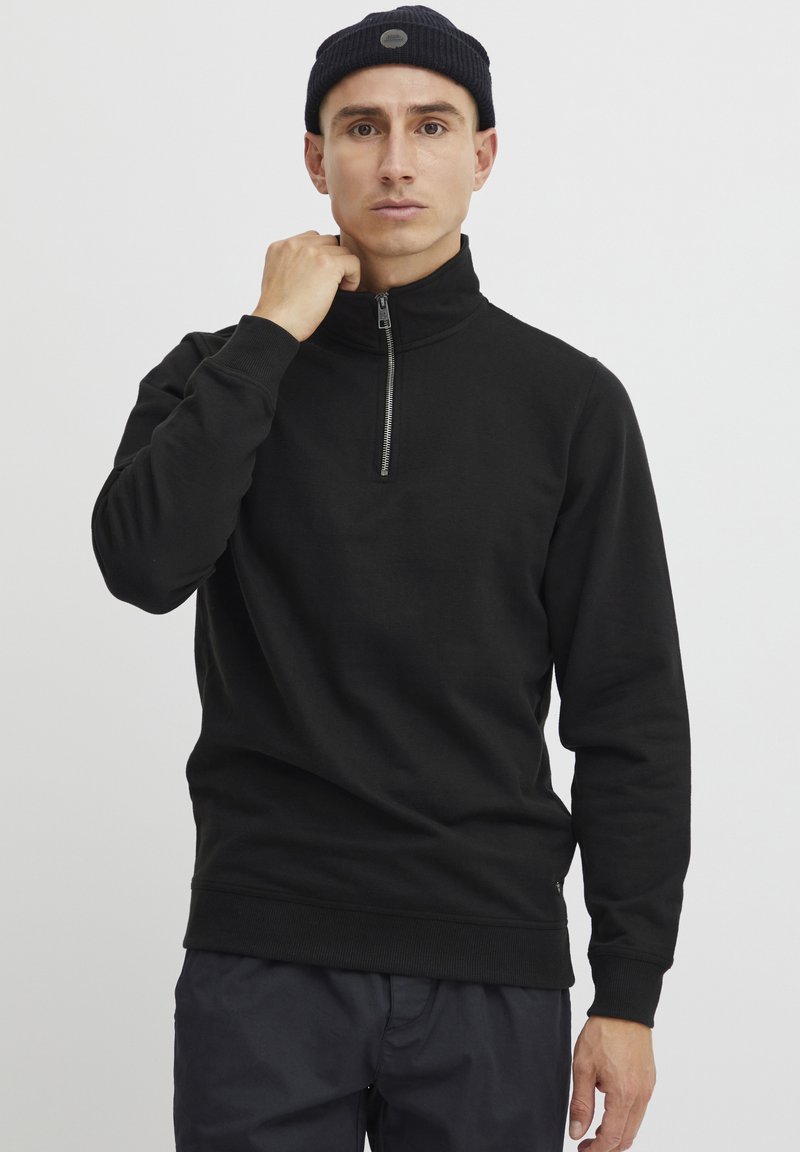 Crna hoodie na zatvarač s visokim ovratnikom, izrađena od glatkog materijala. Ima duge rukave i prilagođeni kroj.