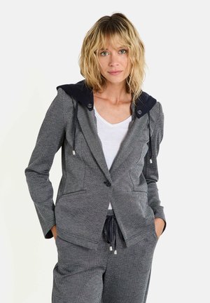 Blazer à motif gris avec une capuche bleu marine, comportant un bouton unique, deux poches avant et des cordons de serrage sur la capuche. Porté par-dessus une chemise blanche.