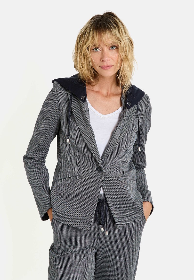 Blazer à motif gris avec une capuche bleu marine, comportant un bouton unique, deux poches avant et des cordons de serrage sur la capuche. Porté par-dessus une chemise blanche.