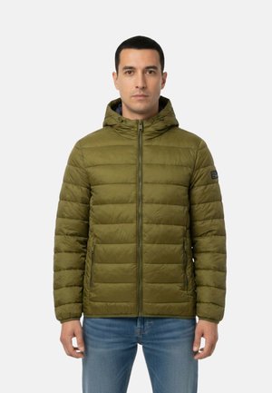 Uomo in piedi di fronte, indossa una giacca imbottita con cappuccio verde oliva e jeans blu su uno sfondo chiaro e uniforme.
