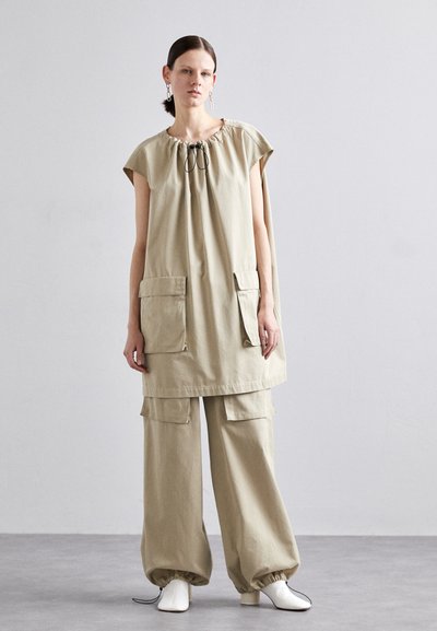 Tunic oversize beige con colletto regolabile, maniche corte e due tasche frontali, abbinato a pantaloni larghi coordinati e scarpe con tacco bianche.