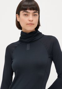 Maglietta nera a maniche lunghe con colletto alto e ripiegato. Presenta un tessuto testurizzato e un piccolo etichetta con logo nella parte anteriore. Design aderente.