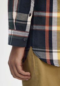 Camicia a quadri con colletto in blu, verde e giallo con polsini a bottone; indossata con pantaloni beige. Presenta un orlo curvo e strisce a contrasto.
