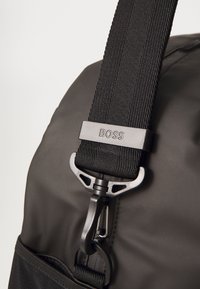 Sort rygsækrem med tykk, struktureret vævet stof og en plastspænde med "BOSS"-branding. Nærbillede af hardware-detaljer.