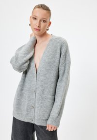 Grå stickad kofta med V-ringning, framknäppning och två fickor. Har ribbad struktur och långa ärmar.