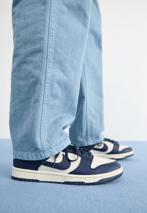Personne portant des baskets basses bleu marine et blanc avec des lacets noirs et un pantalon large bleu clair, debout sur un tapis bleu.