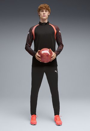 Jonge man in zwart-rode sportkleding die een rode Puma voetbal vasthoudt, staand tegen een effen grijze achtergrond.