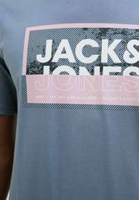 Lichtgrijze katoenen t-shirt met een rechthoekige grafiek. Kenmerkt zich door vetgedrukte zwarte tekst "JACK & JONES" en een roze achtergrond met gestreepte accenten.