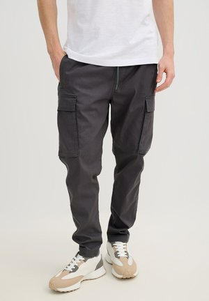 Hombre con pantalones cargo negros con bolsillos delanteros de solapa y zapatillas beige y blancas, de pie sobre un fondo liso.