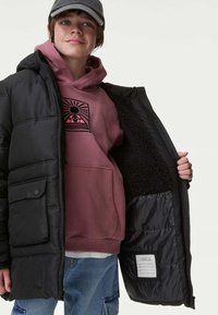 Giacca a vento nera con una tasca frontale, aperta per rivelare una felpa rosa sotto. L'interno è foderato con un morbido peluche nero. Pantaloni in denim casual.