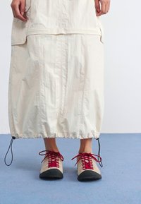 Une personne se tient avec une longue jupe claire cintrée en bas et portant des chaussures beiges avec des lacets rouges sur un sol bleu.